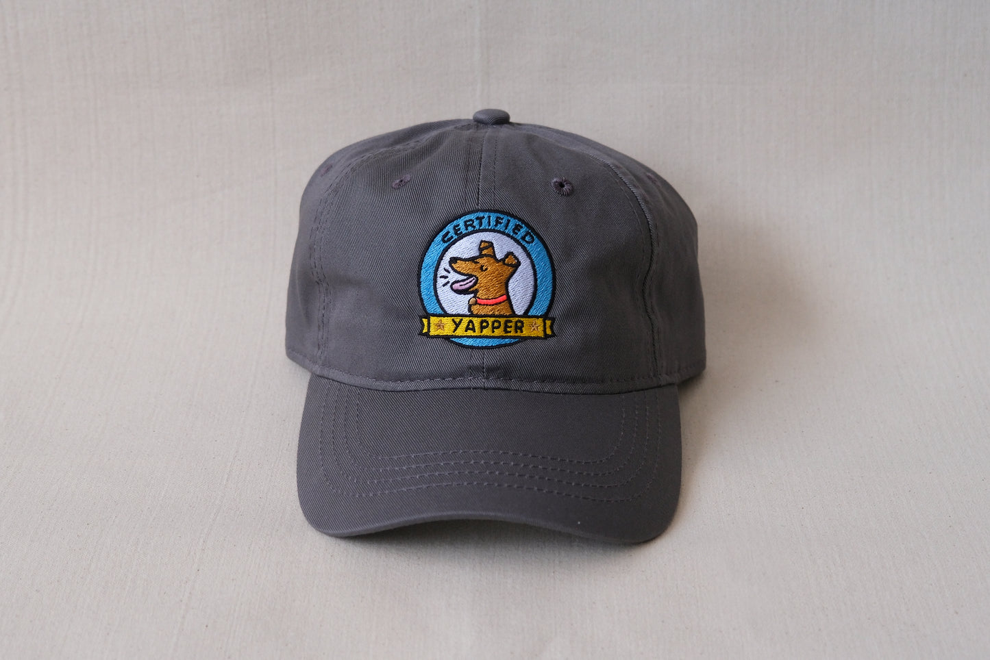 A photo of a grey dad hat with an embroidered dog that says "Certified Yapper" over a cream background.