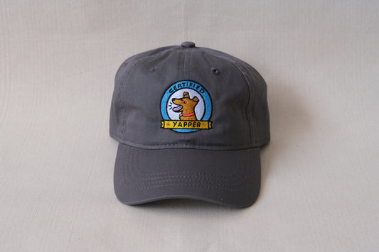 A photo of a grey dad hat with an embroidered dog that says "Certified Yapper" over a cream background.