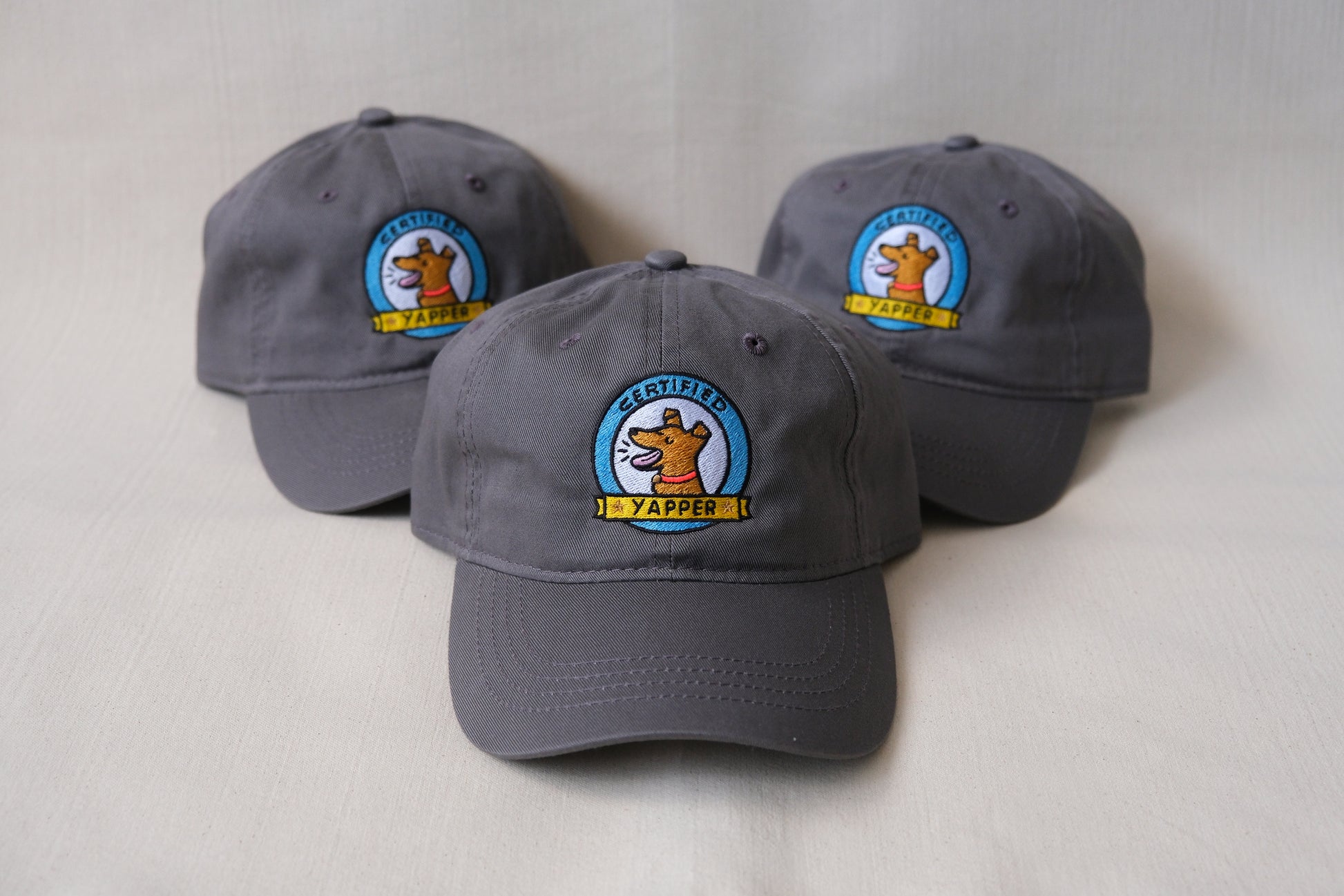 A photo of a 3 grey dad hats embroidered with dogs that say "Certified Yapper" over a cream background.
