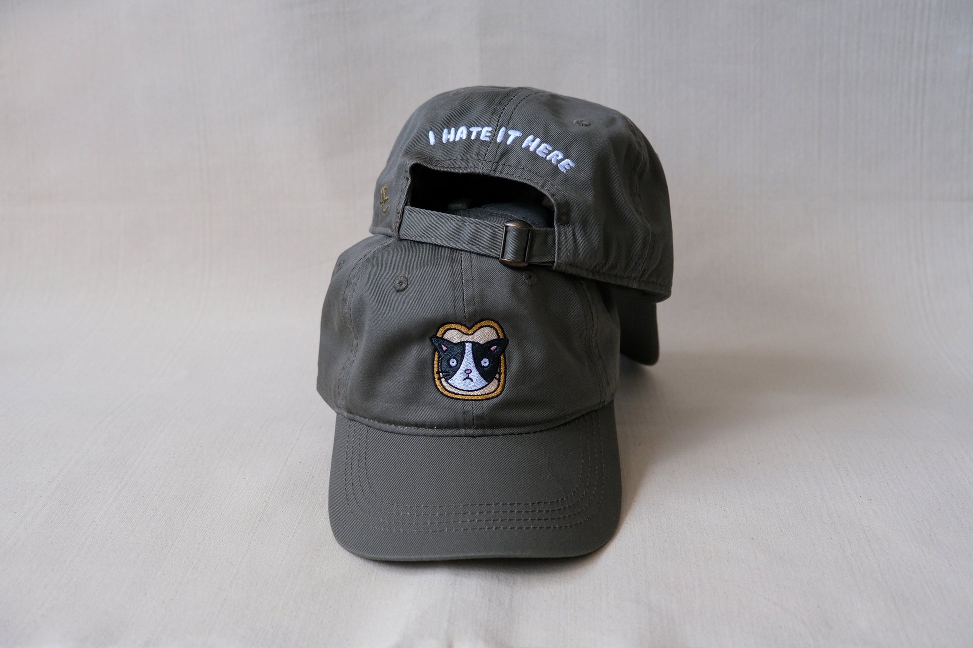 A photo of an olive green dad hat with an embroidered cat stuck in a slice of bread and the back of a second hat with "I Hate It Here" embroidered on the back in white.