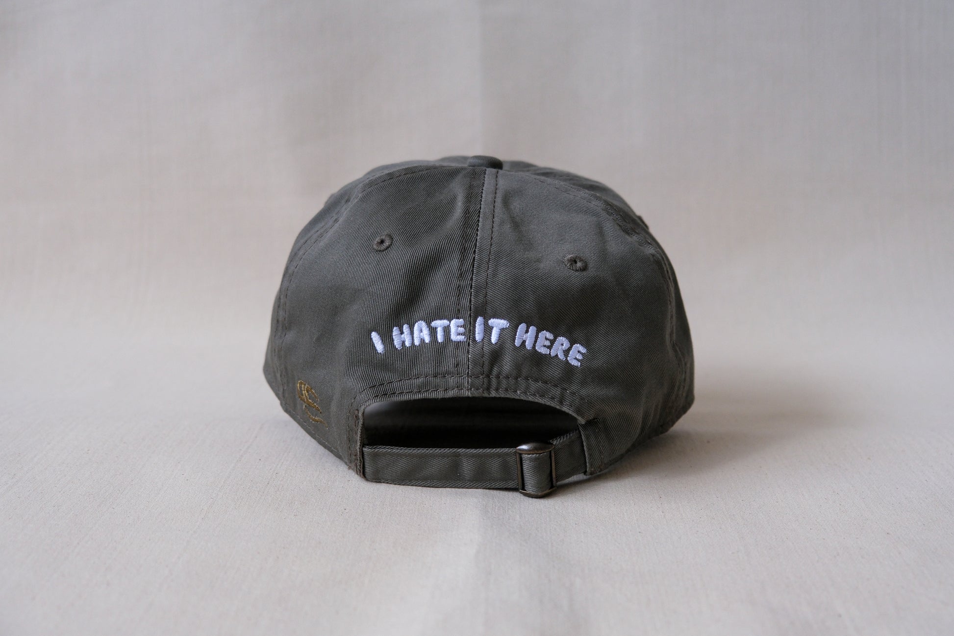 A photo of the back of an olive green dad hat that says "I Hate It Here" embroidered in white with an adjustable buckle strap.
