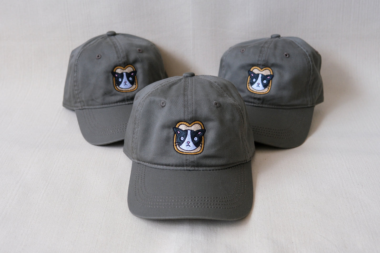 A photo of 3 olive green dad hats with embroidered cats stuck in a slices of bread over a cream background.