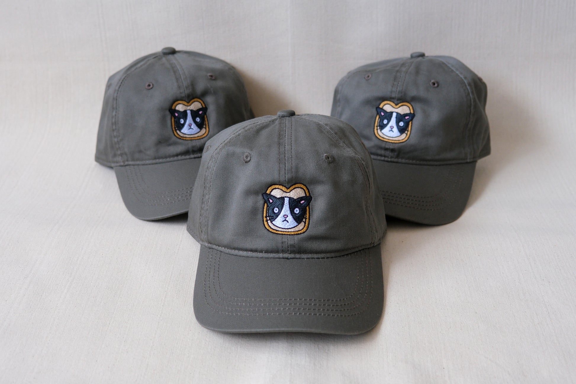 A photo of 3 olive green dad hats with embroidered cats stuck in a slices of bread over a cream background.