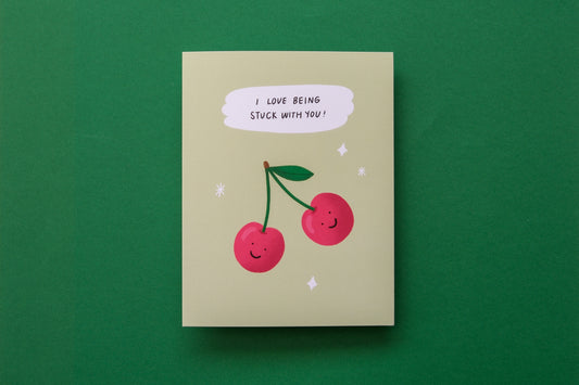 A photo of a greeting card with 2 cherries that says "I love being stuck with you" on a green background.