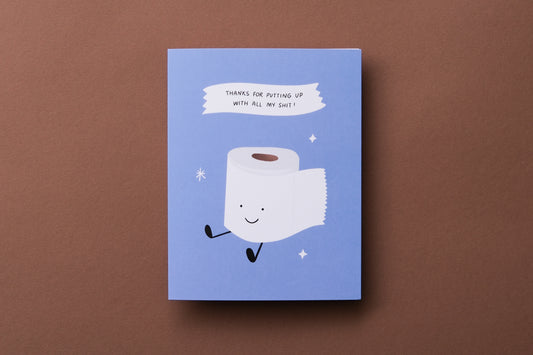 A photo of a greeting card with a smiling roll of toilet paper that says "Thanks for putting up with all my shit!" and a white envelope on a brown background.