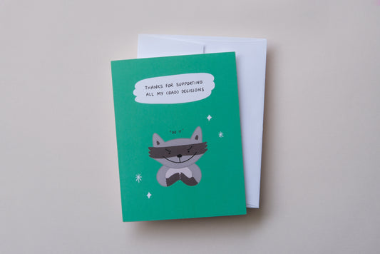 A photo of a greeting card with an evil raccoon that says "Thanks for supporting all my (bad) decisions" on a cream background.
