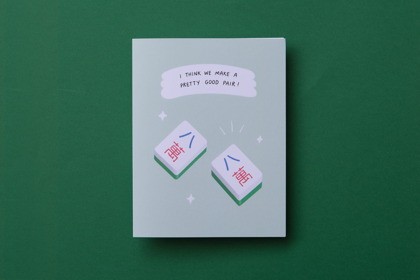 A photo of a greeting card with a pair of matching mahjong tiles "I think we make a pretty good pair!" and a white envelope on a green background.
