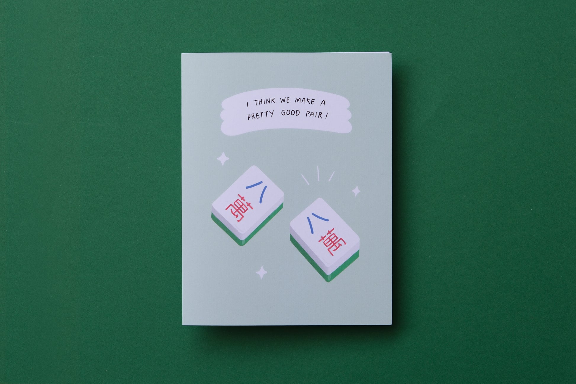 A photo of a greeting card with a pair of matching mahjong tiles "I think we make a pretty good pair!" and a white envelope on a green background.