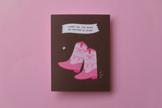 A photo of a greeting card with a pair of pink cowboy boots that says "Thank you for being my partner in crime" and a white envelope on a pink background.