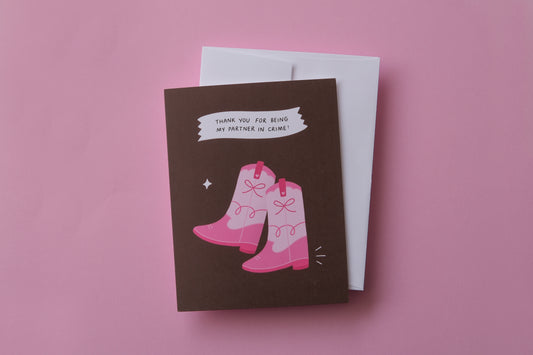 A photo of a greeting card with a pair of pink cowboy boots that says "Thank you for being my partner in crime" on a pink background.