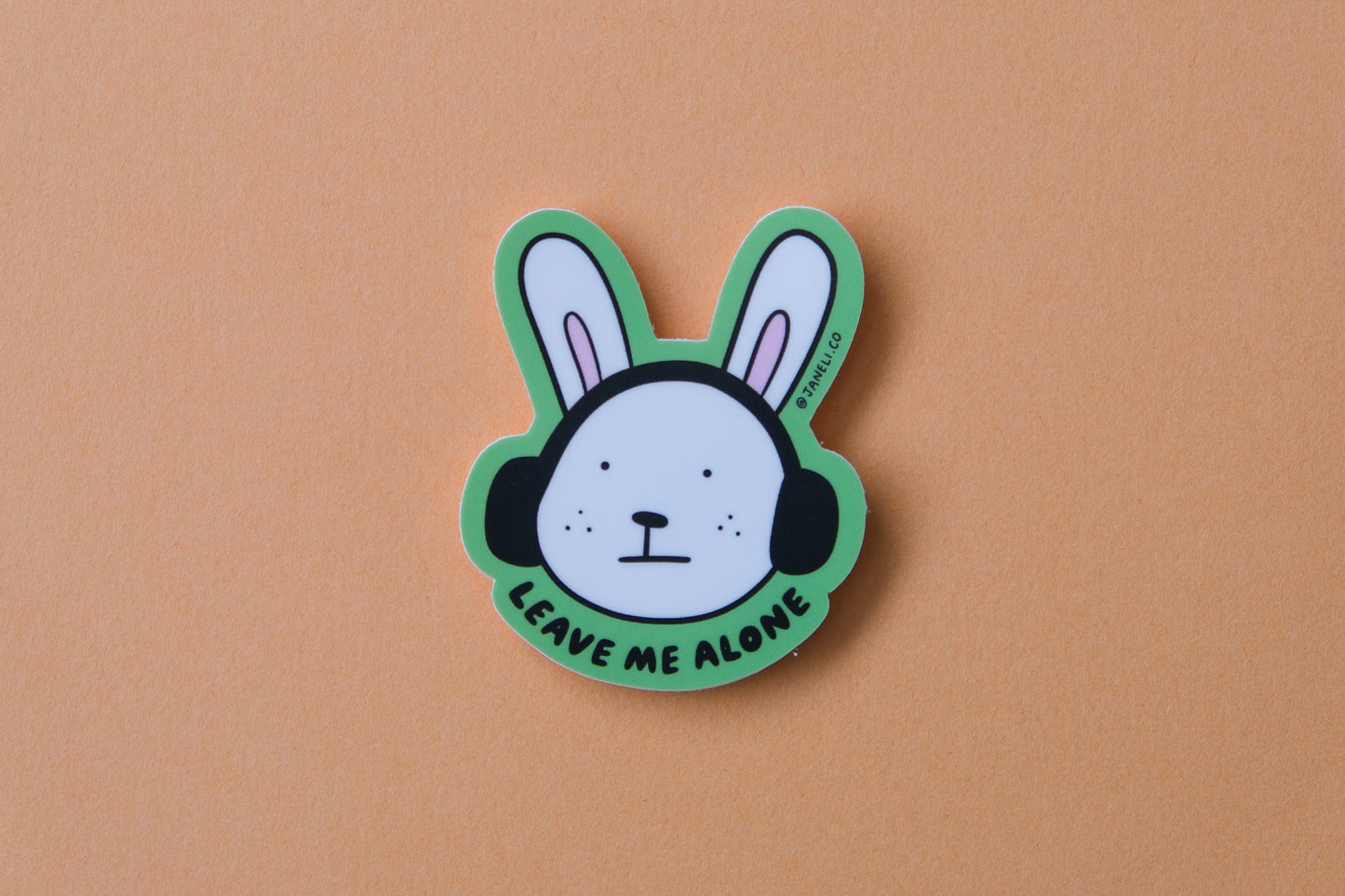 A JaneLi.Co sticker of a bunny with headphones on that says "Leave me alone" over an orange background.