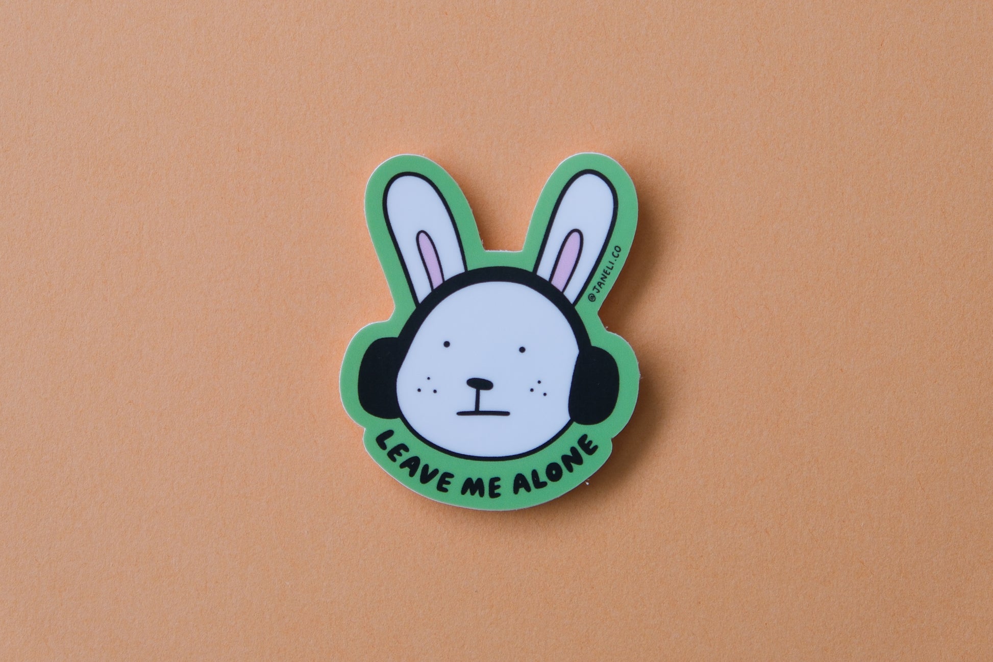 A JaneLi.Co sticker of a bunny with headphones on that says "Leave me alone" over an orange background.
