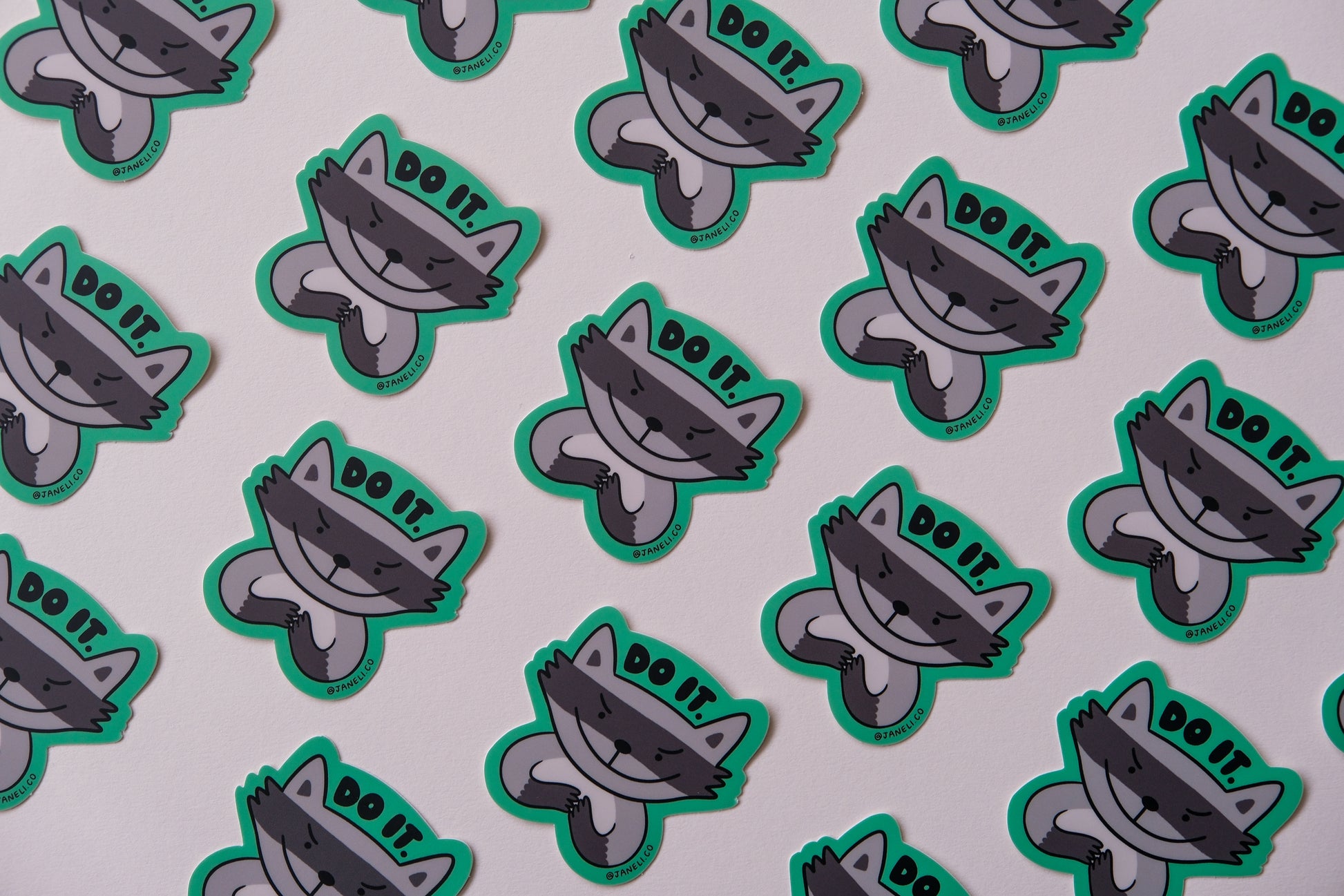 A grid of JaneLi.Co stickers of evil raccoons that say "Do it" over an cream background.