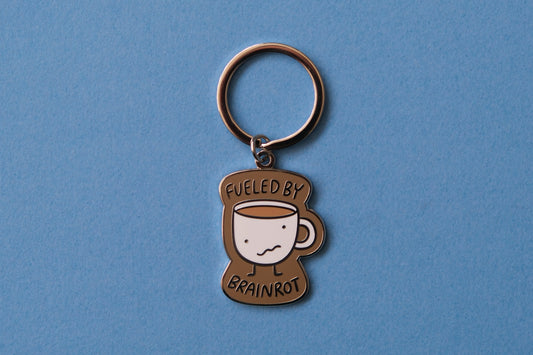 An enamel keychain of a wonky coffee mug that says "Fueled by brainrot" over a blue background.