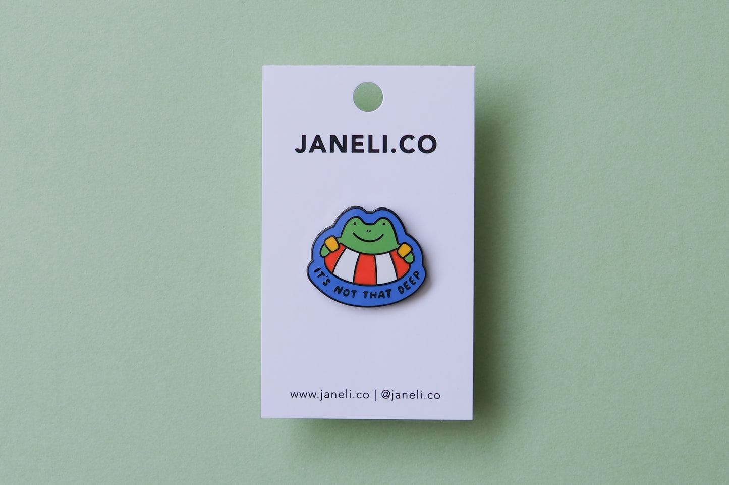 An enamel pin of a frog floating in a lifessaver tube that says "It's not that deep" over a green background.