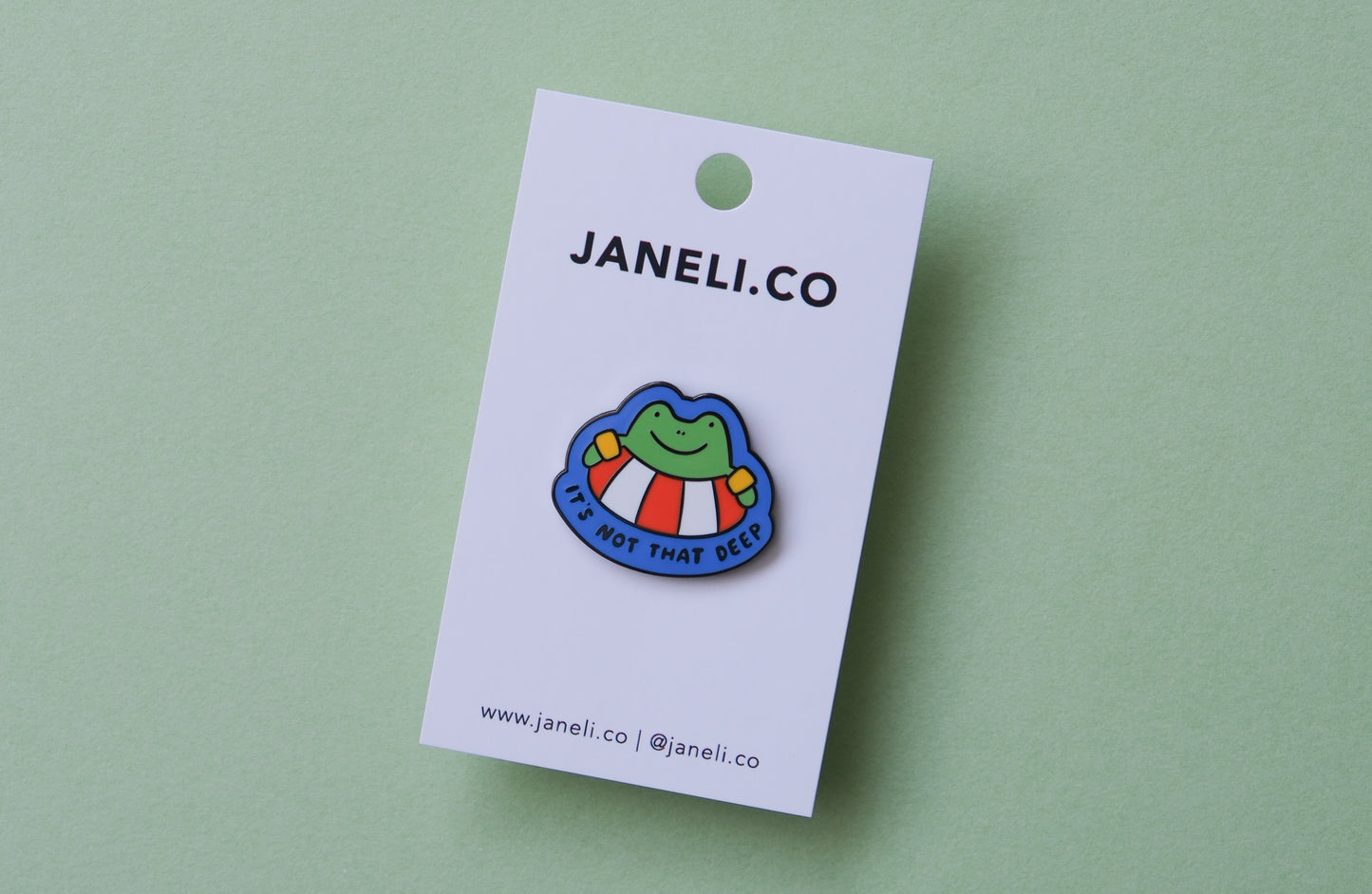 An enamel pin of a frog floating in a lifessaver tube that says "It's not that deep" over a green background.