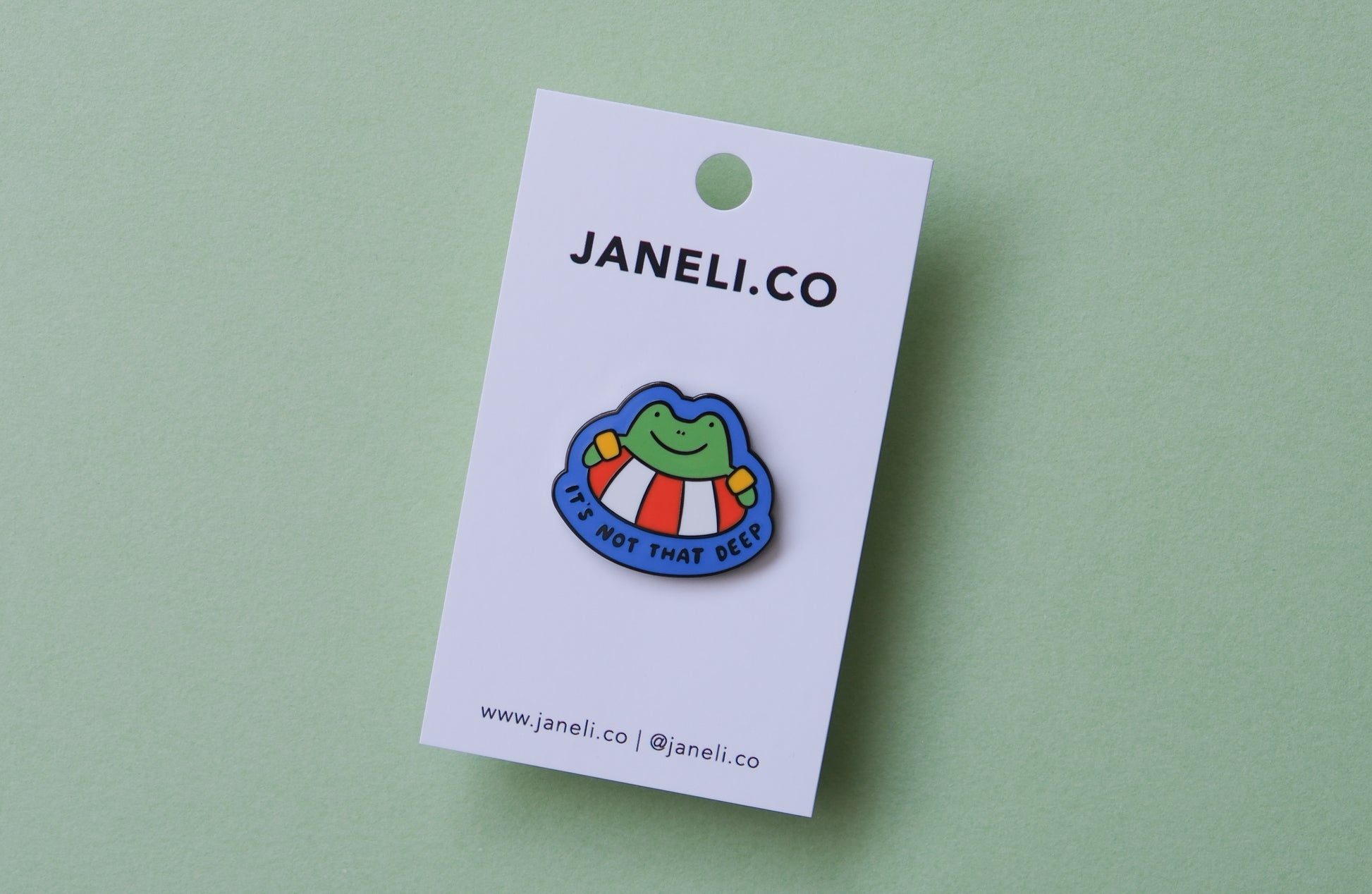 An enamel pin of a frog floating in a lifessaver tube that says "It's not that deep" over a green background.