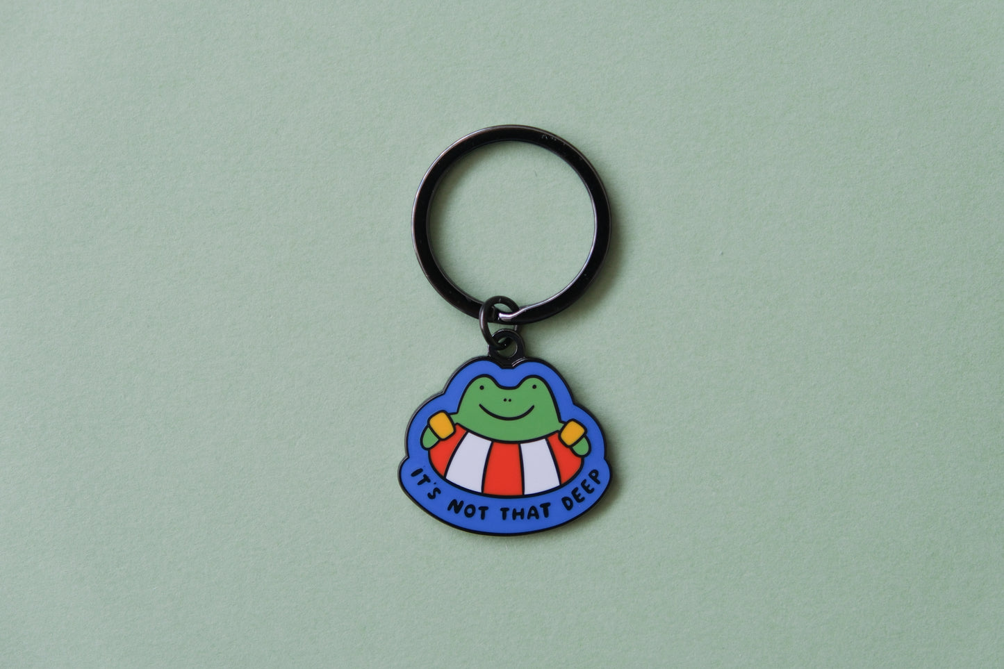An enamel keychain of a frog floating in a lifessaver tube that says "It's not that deep" over a green background.