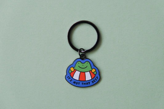 An enamel keychain of a frog floating in a lifessaver tube that says "It's not that deep" over a green background.
