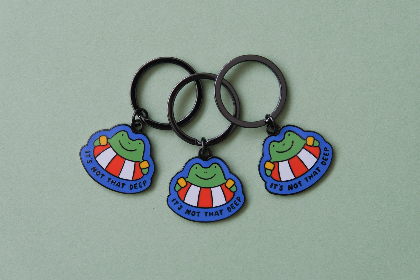 3 enamel keychains of frogs floating in lifessaver tubes that say "It's not that deep" over a green background.