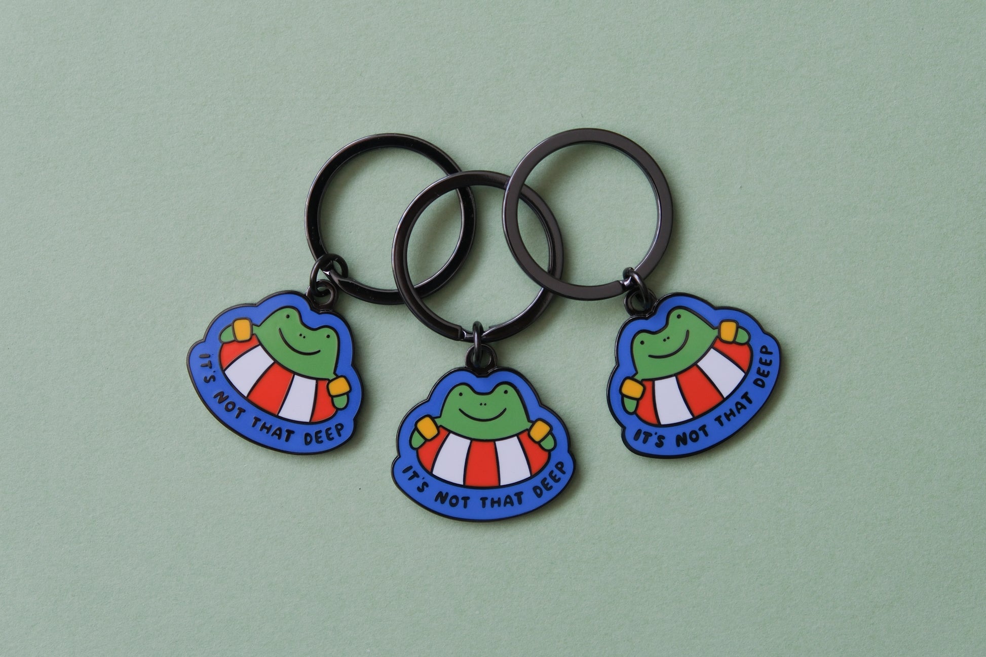 3 enamel keychains of frogs floating in lifessaver tubes that say "It's not that deep" over a green background.