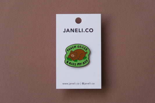 An enamel pin of a napping brown bear that says "Touch grass and kiss my ass" over a brown background.