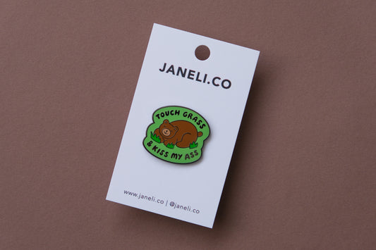 An enamel pin of a napping brown bear that says "Touch grass and kiss my ass" over a brown background.