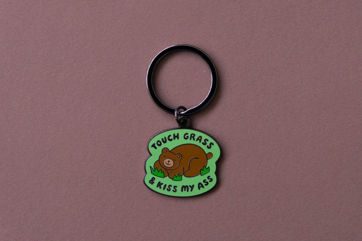 An enamel keychain of a napping brown bear that says "Touch grass and kiss my ass" over a brown background.
