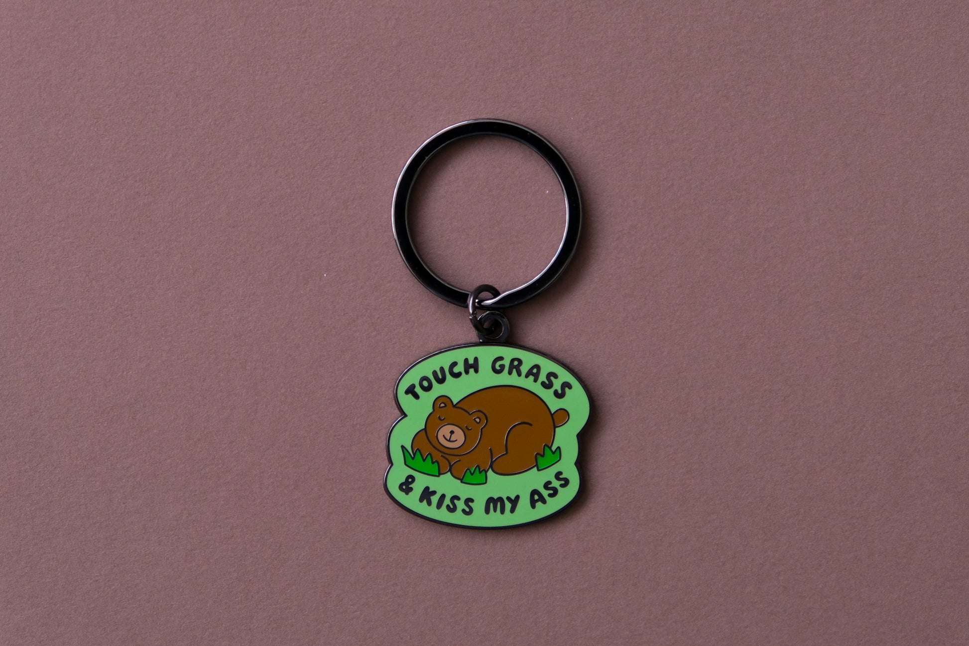 An enamel keychain of a napping brown bear that says "Touch grass and kiss my ass" over a brown background.