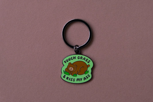 An enamel keychain of a napping brown bear that says "Touch grass and kiss my ass" over a brown background.