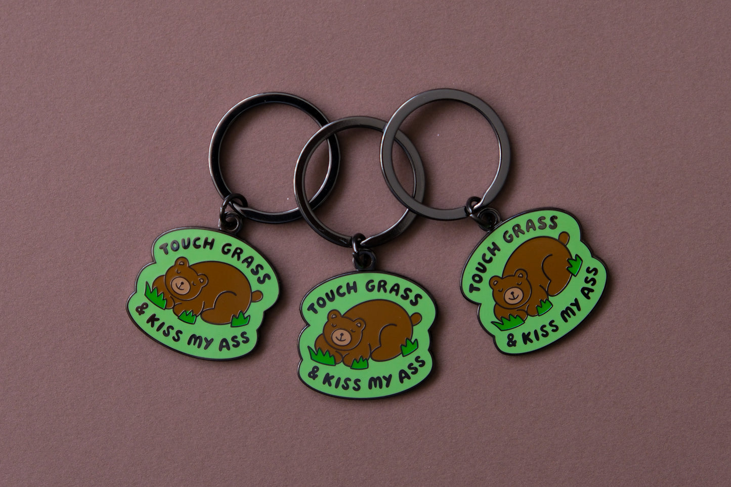 3 enamel keychains of napping brown bears that say "Touch grass and kiss my ass" over a brown background.