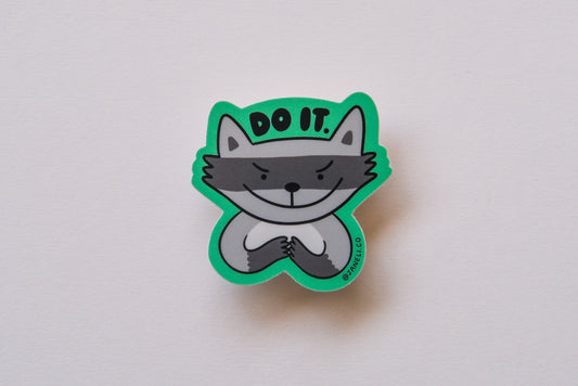 A JaneLi.Co sticker of an evil raccoon that says "Do it" over an cream background.