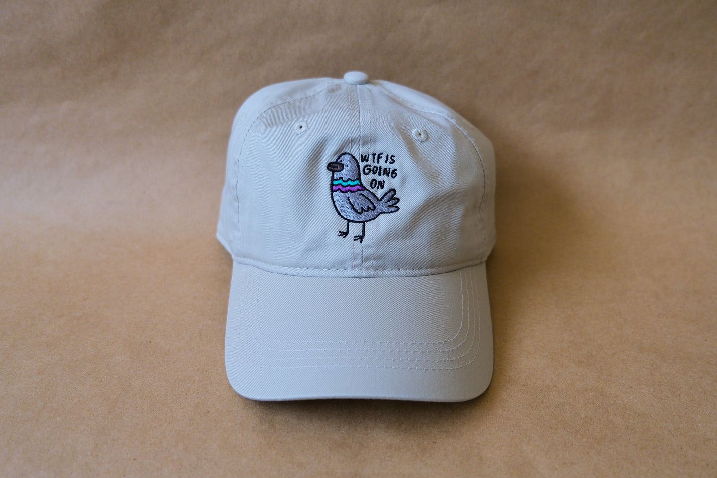 A photo of a khaki dad hat with an embroidered pigeon that says "WTF Is Going On" over a tan background.