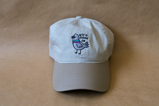A photo of a khaki dad hat with an embroidered pigeon that says "WTF Is Going On" over a tan background.