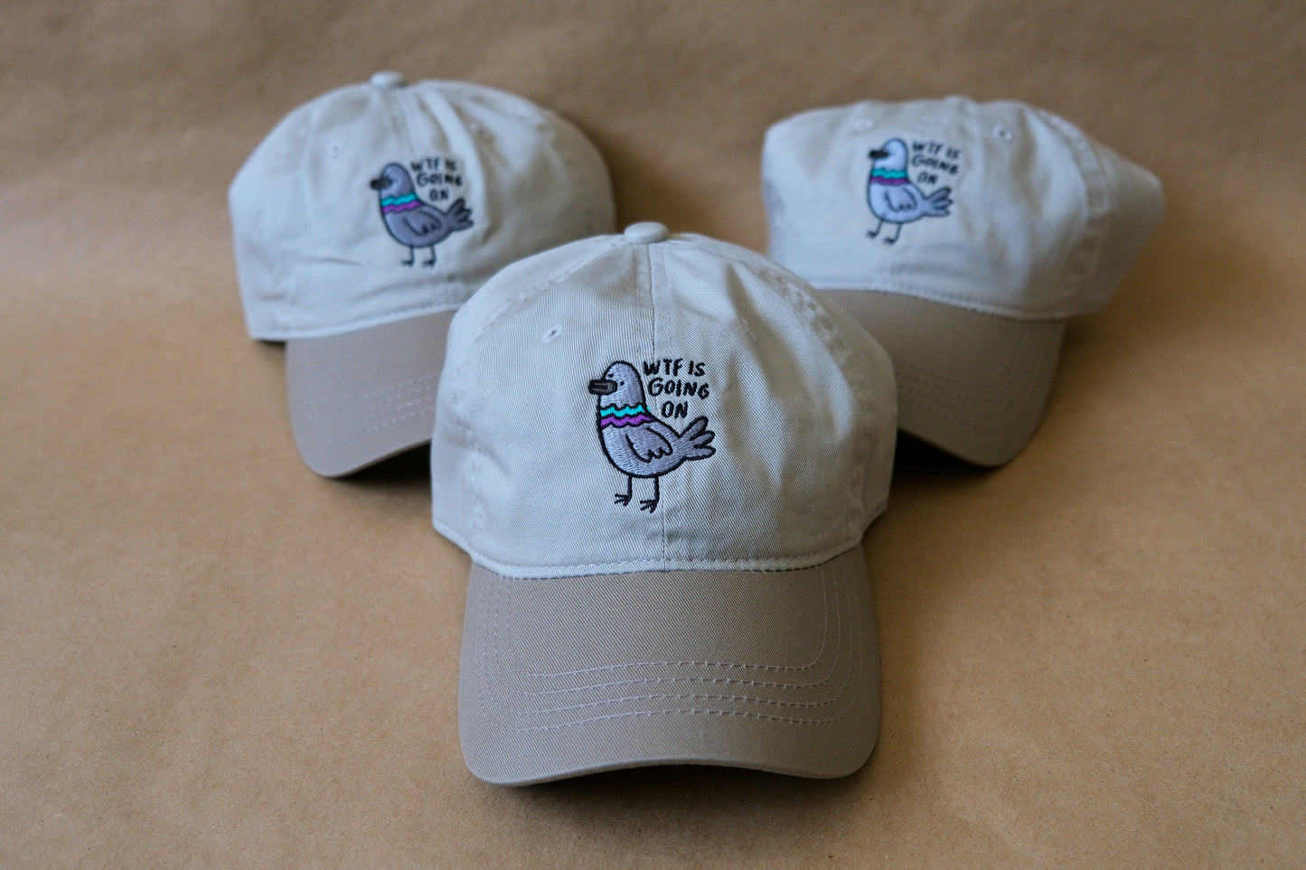 A photo of 3 khaki dad hats with embroidered pigeons that say "WTF Is Going On" over a tan background.