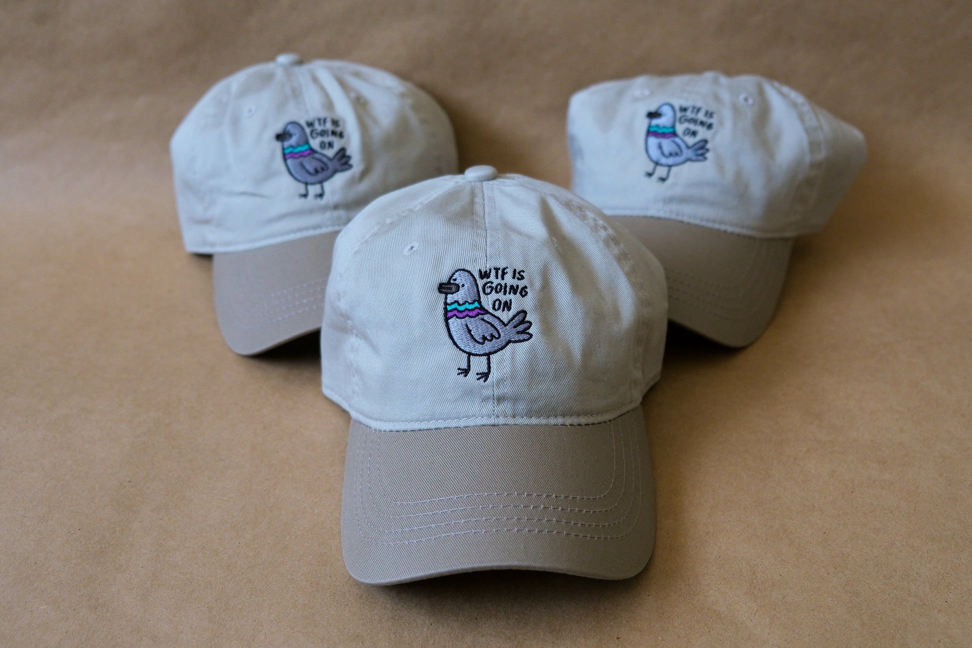 A photo of 3 khaki dad hats with embroidered pigeons that say "WTF Is Going On" over a tan background.