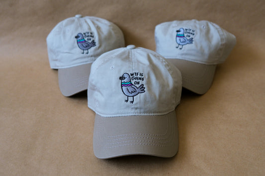 A photo of 3 khaki dad hats with embroidered pigeons that say "WTF Is Going On" over a tan background.