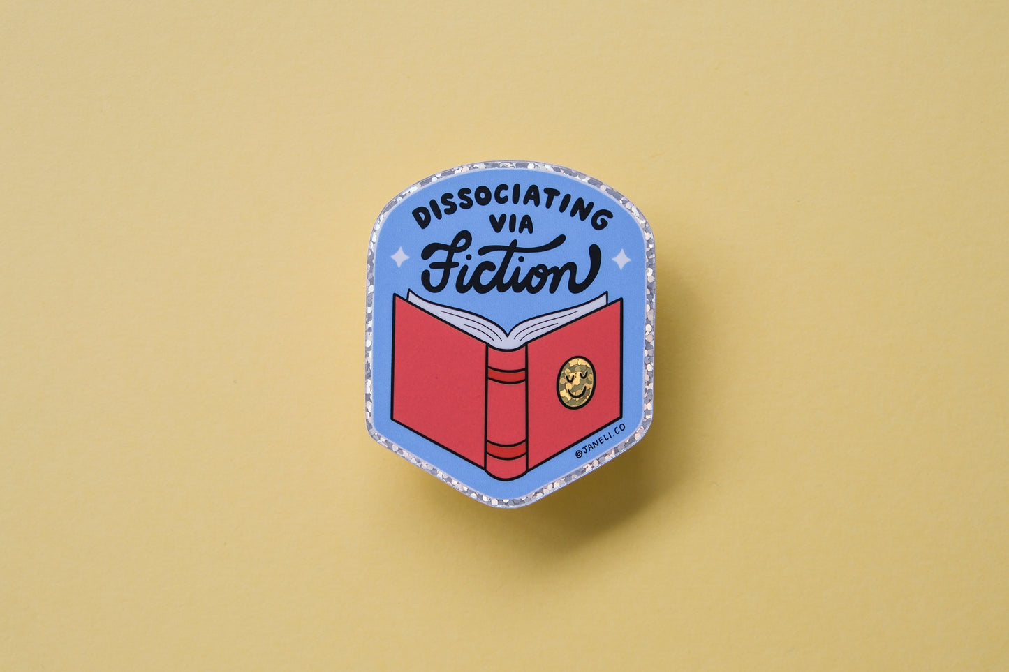 A JaneLi.Co sticker of badge with a book that says "Dissociating via Fiction" over a dark yellow background.