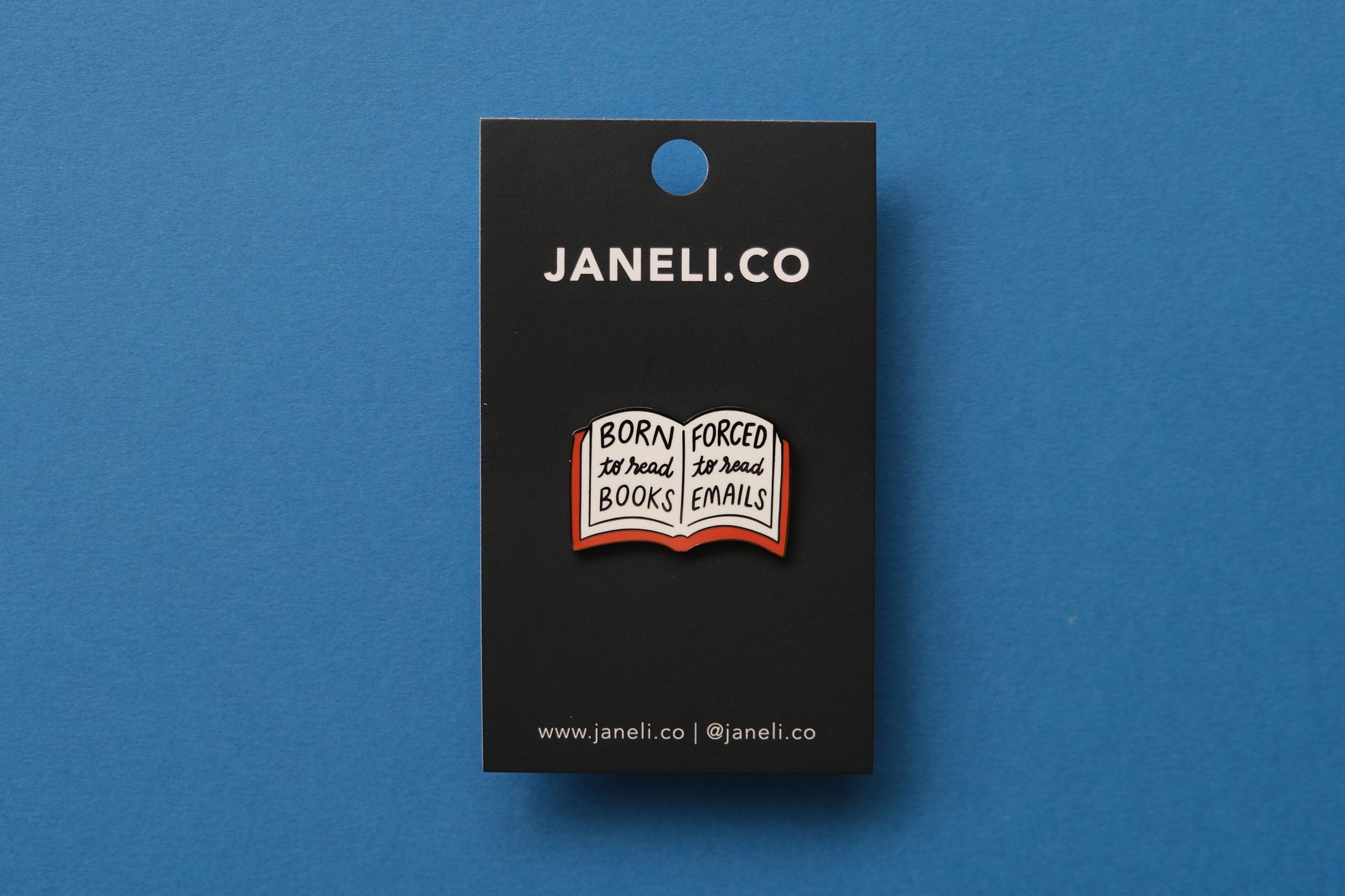 An enamel pin of an open book that says "Born to read books, Forced to read emails" over a blue background.