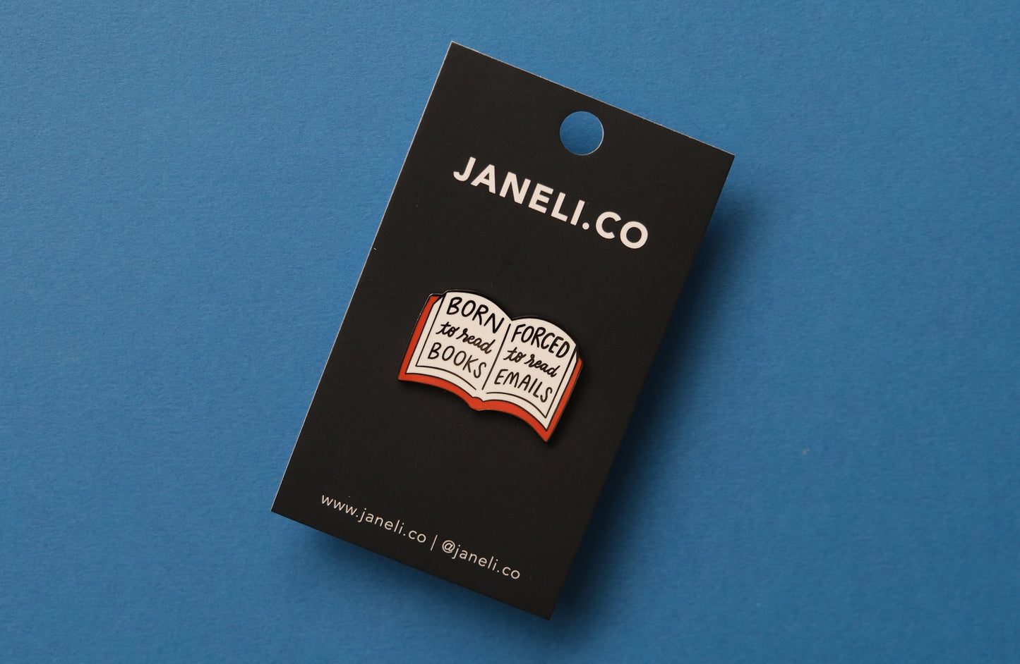 An enamel pin of an open book that says "Born to read books, Forced to read emails" over a blue background.