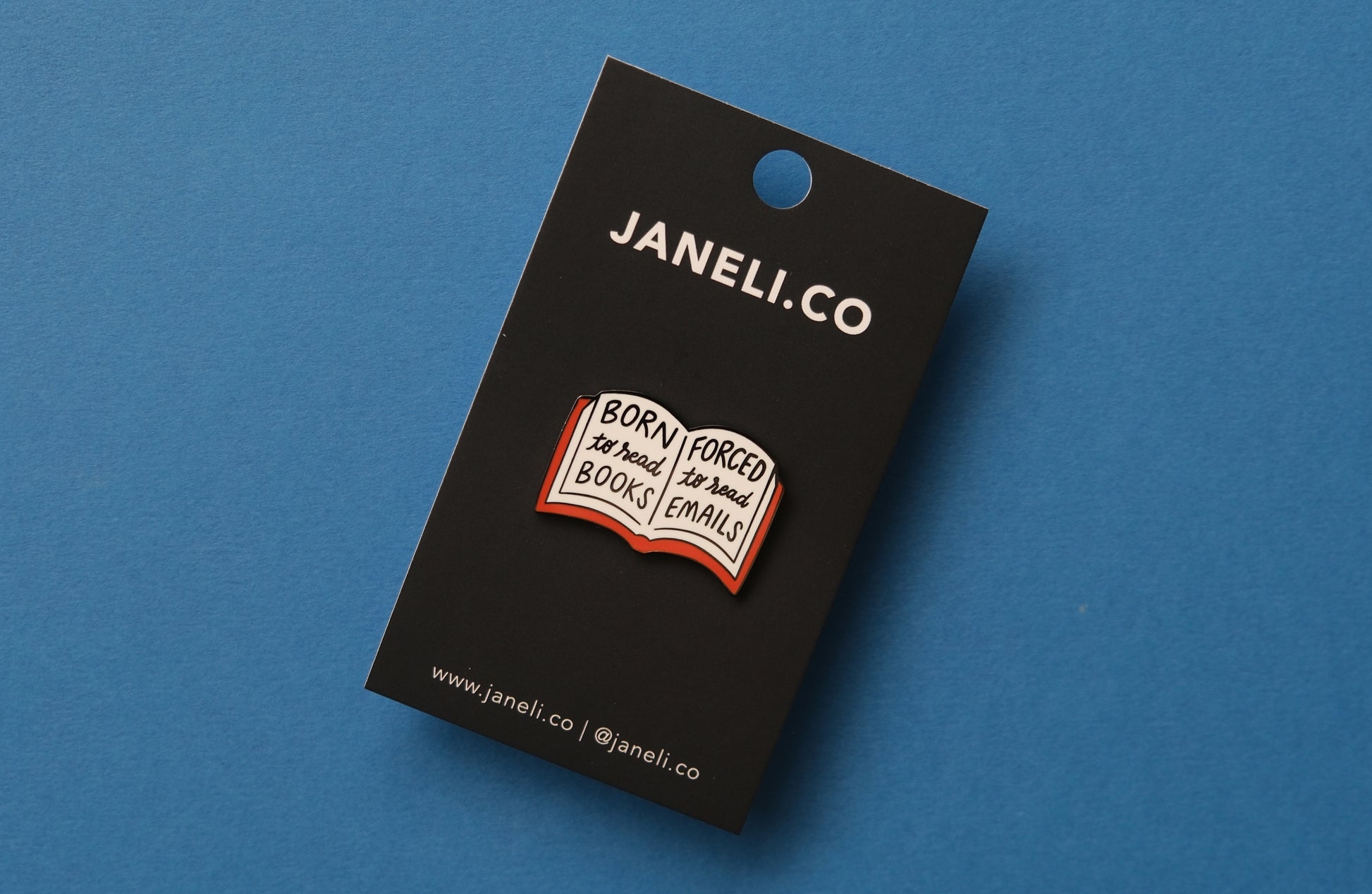 An enamel pin of an open book that says "Born to read books, Forced to read emails" over a blue background.