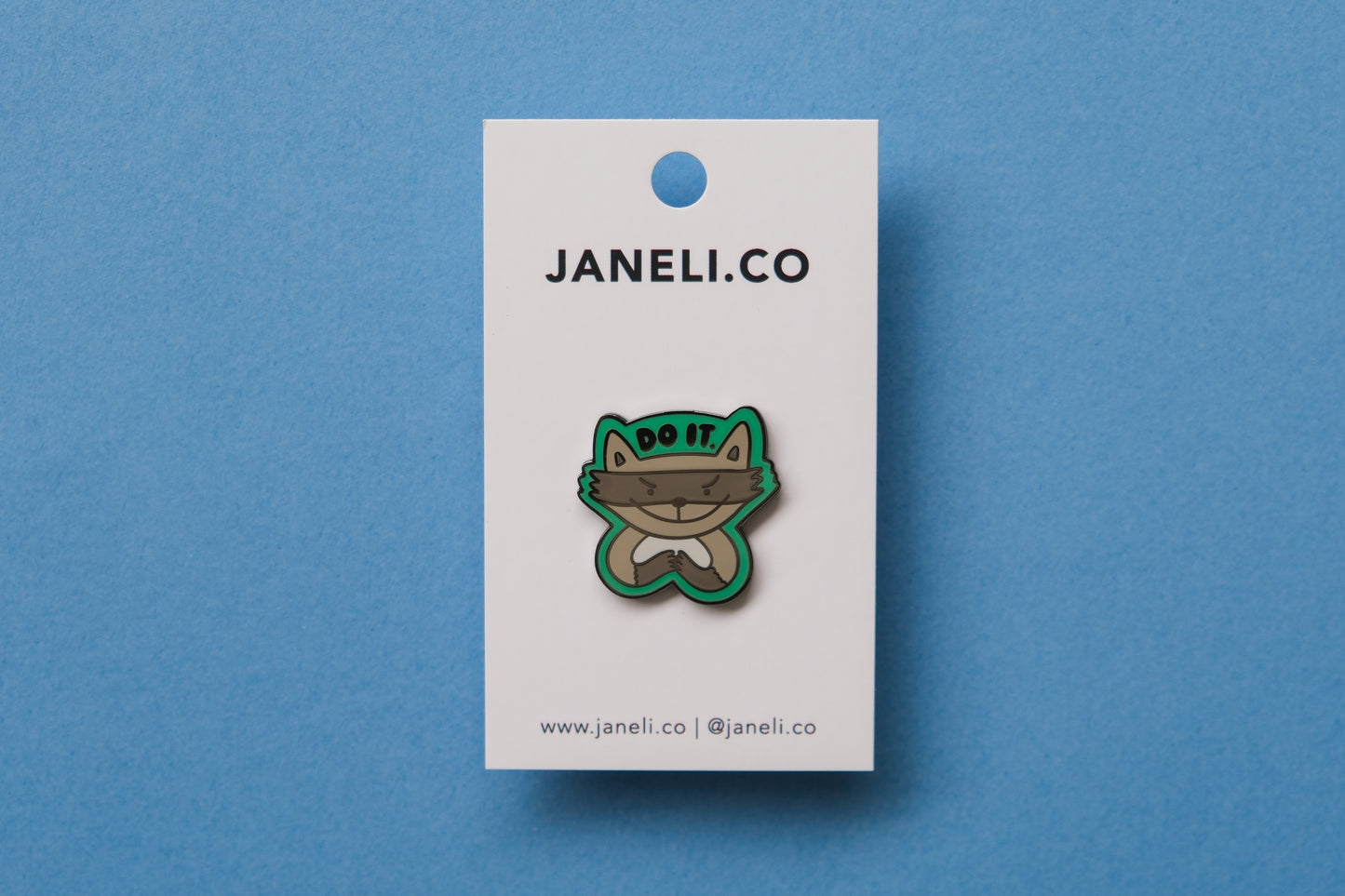 An enamel pin of an evil raccoon that says "Do it." over a blue background.