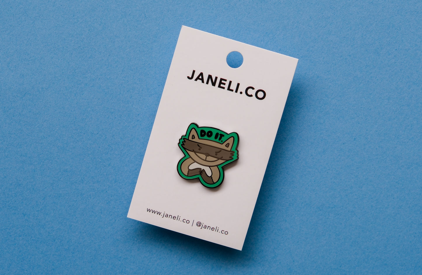 An enamel pin of an evil raccoon that says "Do it." over a blue background.