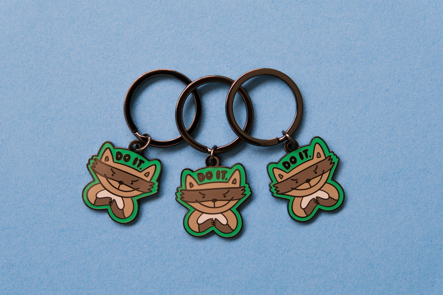 3 enamel keychains of evil raccoons that say "Do it." over a blue background.