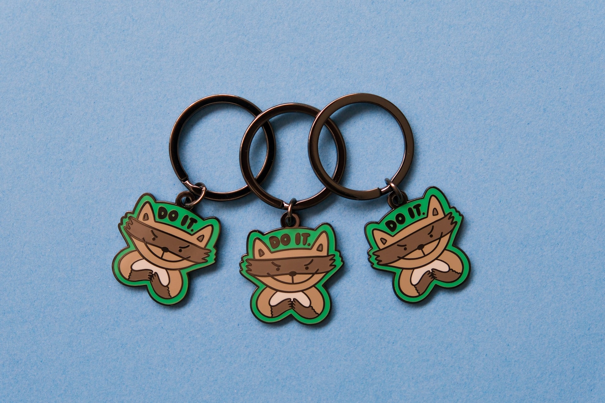 3 enamel keychains of evil raccoons that say "Do it." over a blue background.