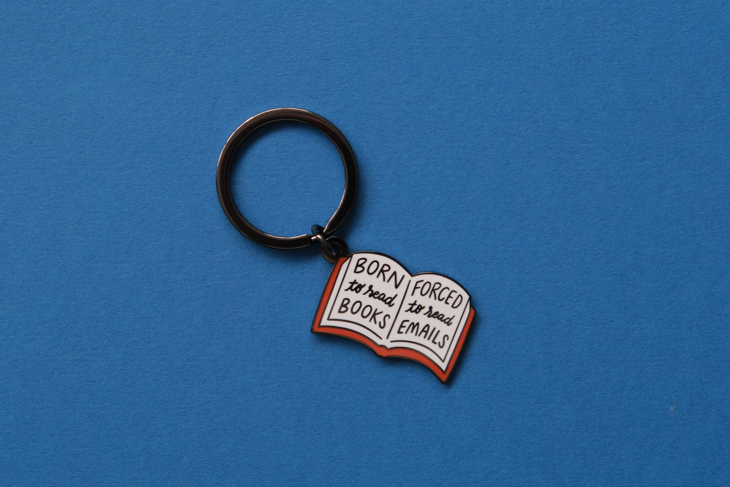 An enamel keychain of an open book that says "Born to read books, Forced to read emails" over a blue background.
