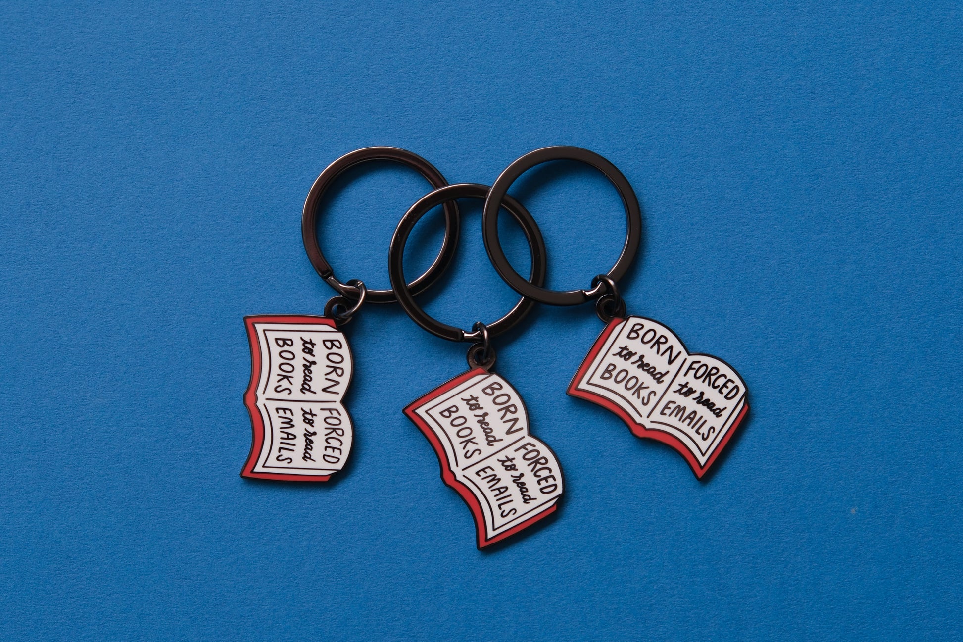 3 enamel keychains of open books that say "Born to read books, Forced to read emails" over a blue background.