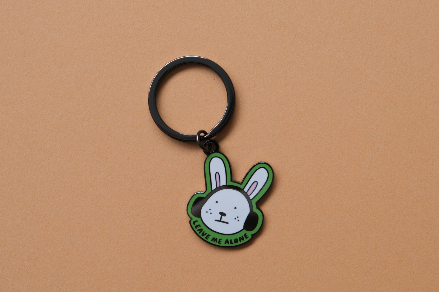 An enamel keychain of a rabbit wearing headphones that says "Leave me alone" over an orange background.