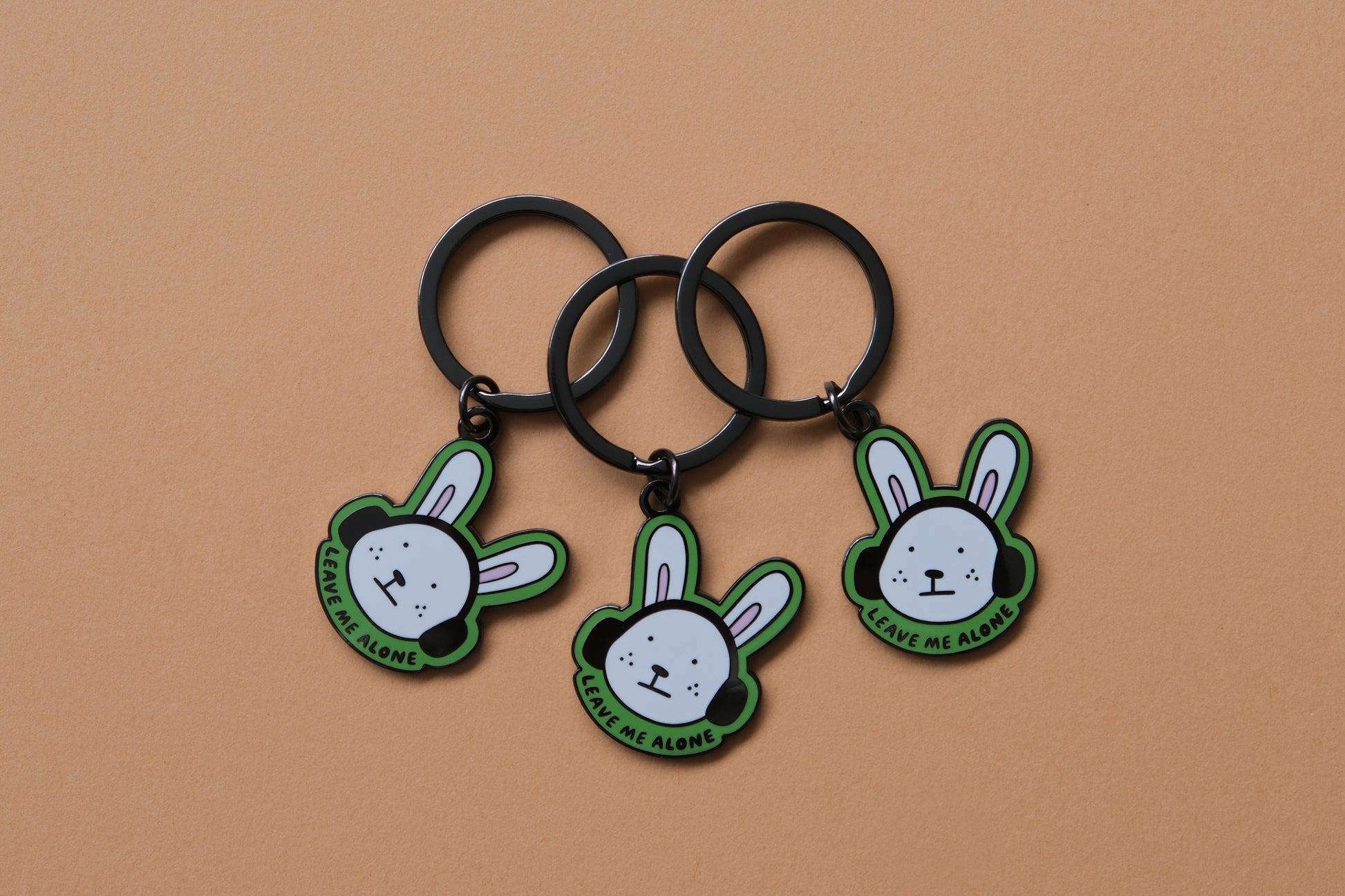 3 enamel keychains of rabbits wearing headphones that say "Leave me alone" over an orange background.