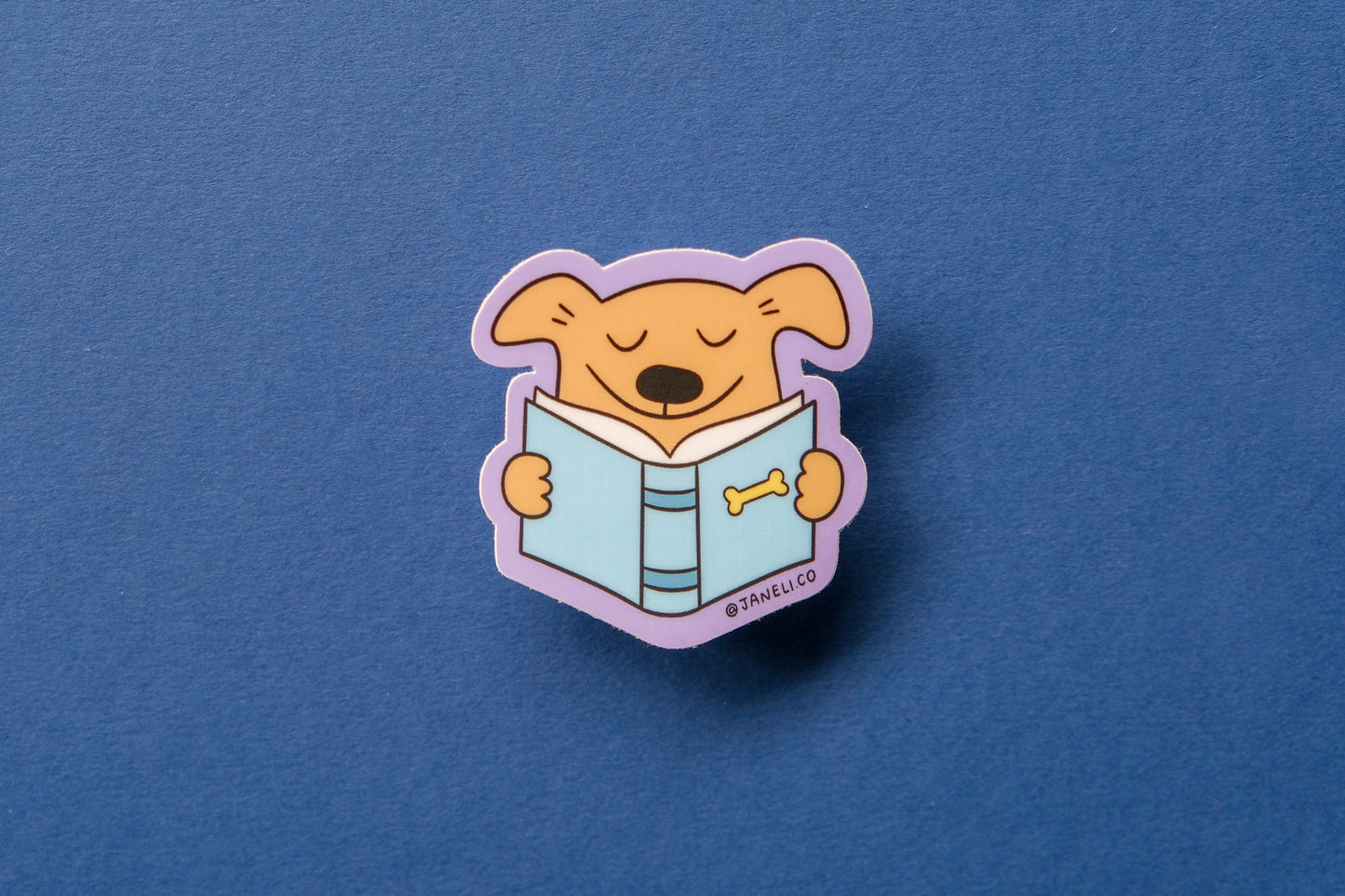 A JaneLi.Co sticker of a dog reading a book over a dark blue background.